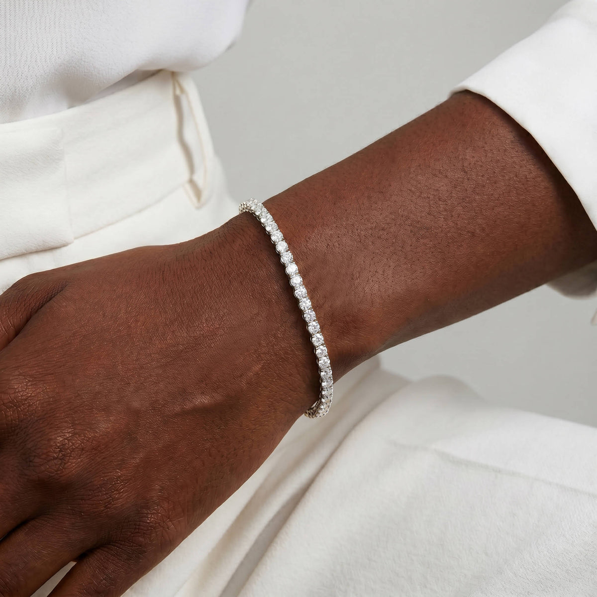A close-up of a person's wrist wearing a delicate silver tennis bracelet, paired with a white top and white pants.