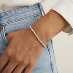 A delicate silver tennis bracelet with clear round-cut stones worn on a woman's wrist, paired with light wash blue jeans and a white top.