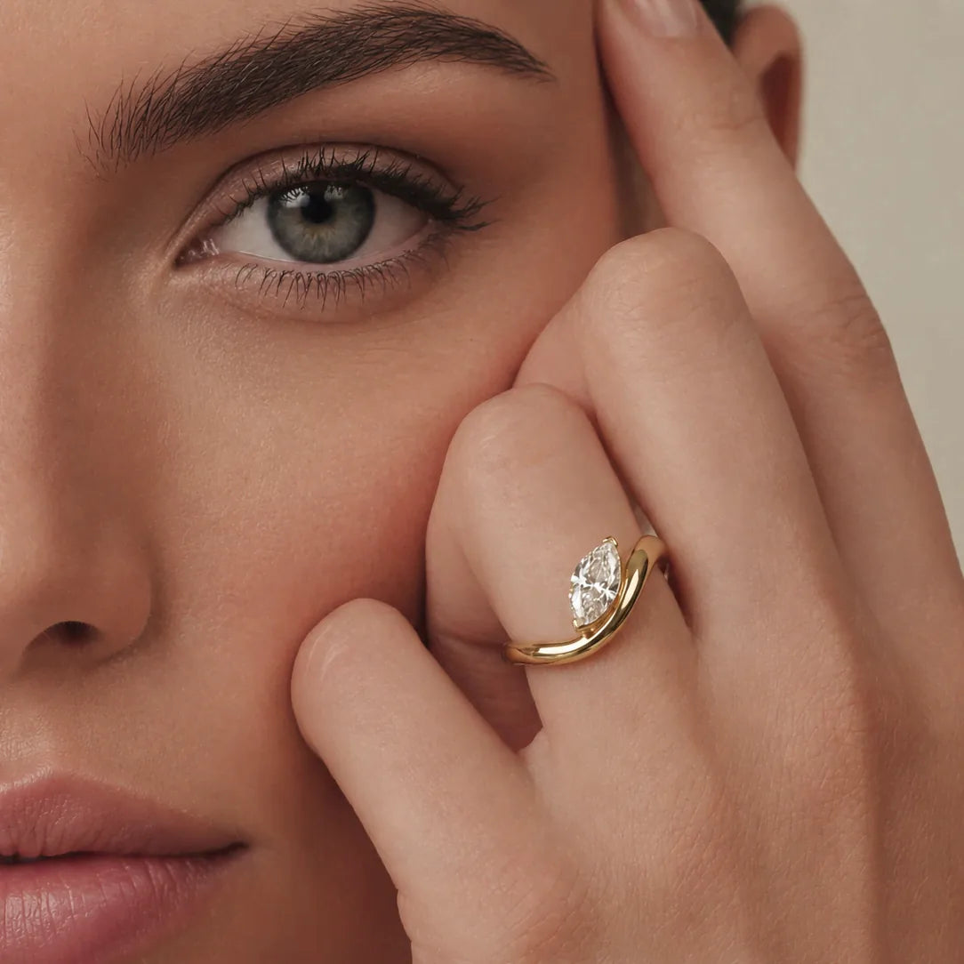 A close-up lifestyle shot of a model wearing a gold ring with a marquise-cut diamond on her finger. The ring features a curved gold band with a central marquise-cut diamond set in a bezel.